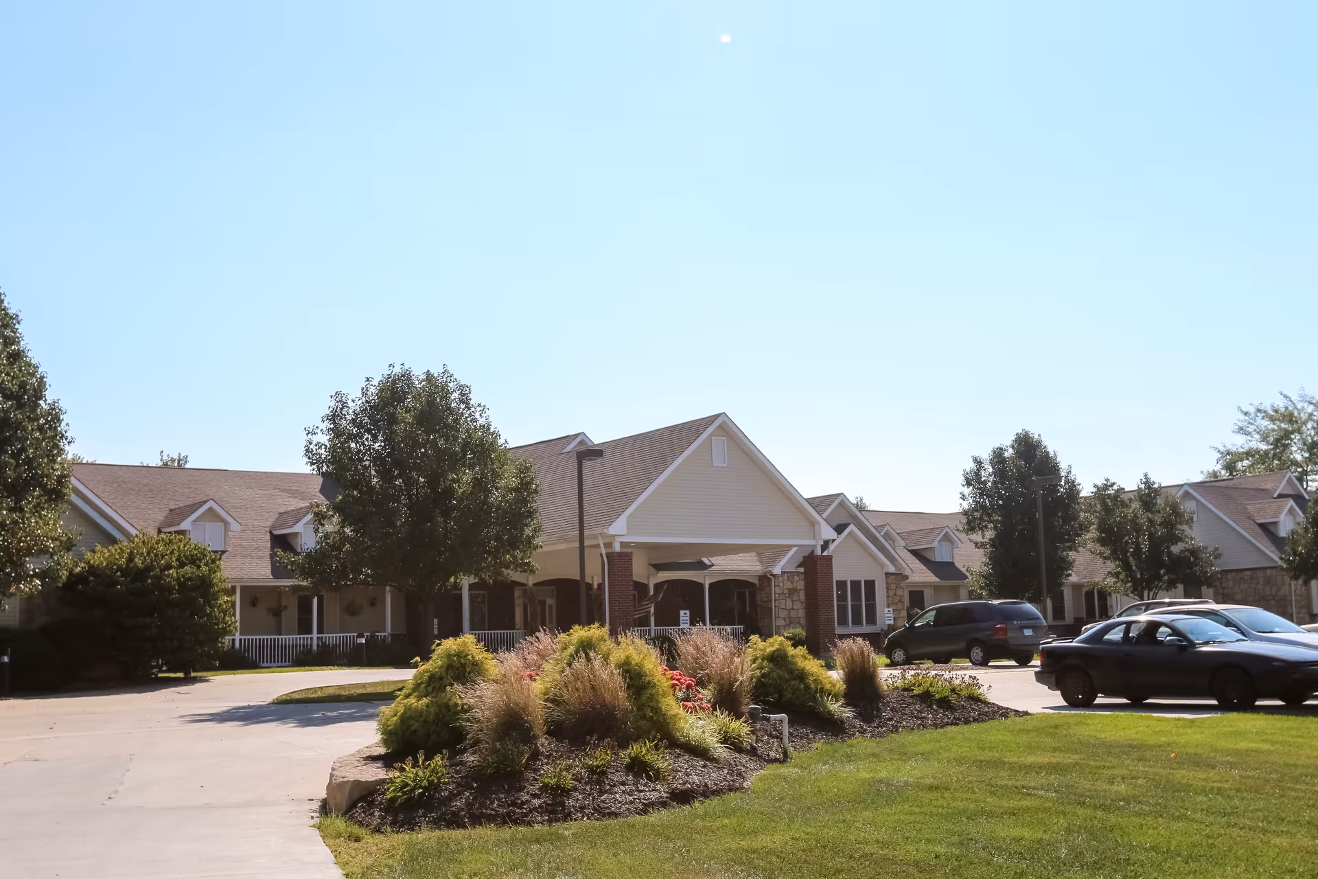 Exterior view of a senior living facility with a driveway, landscaped garden with bushes and flowers, several trees, and parked cars under a clear blue sky.