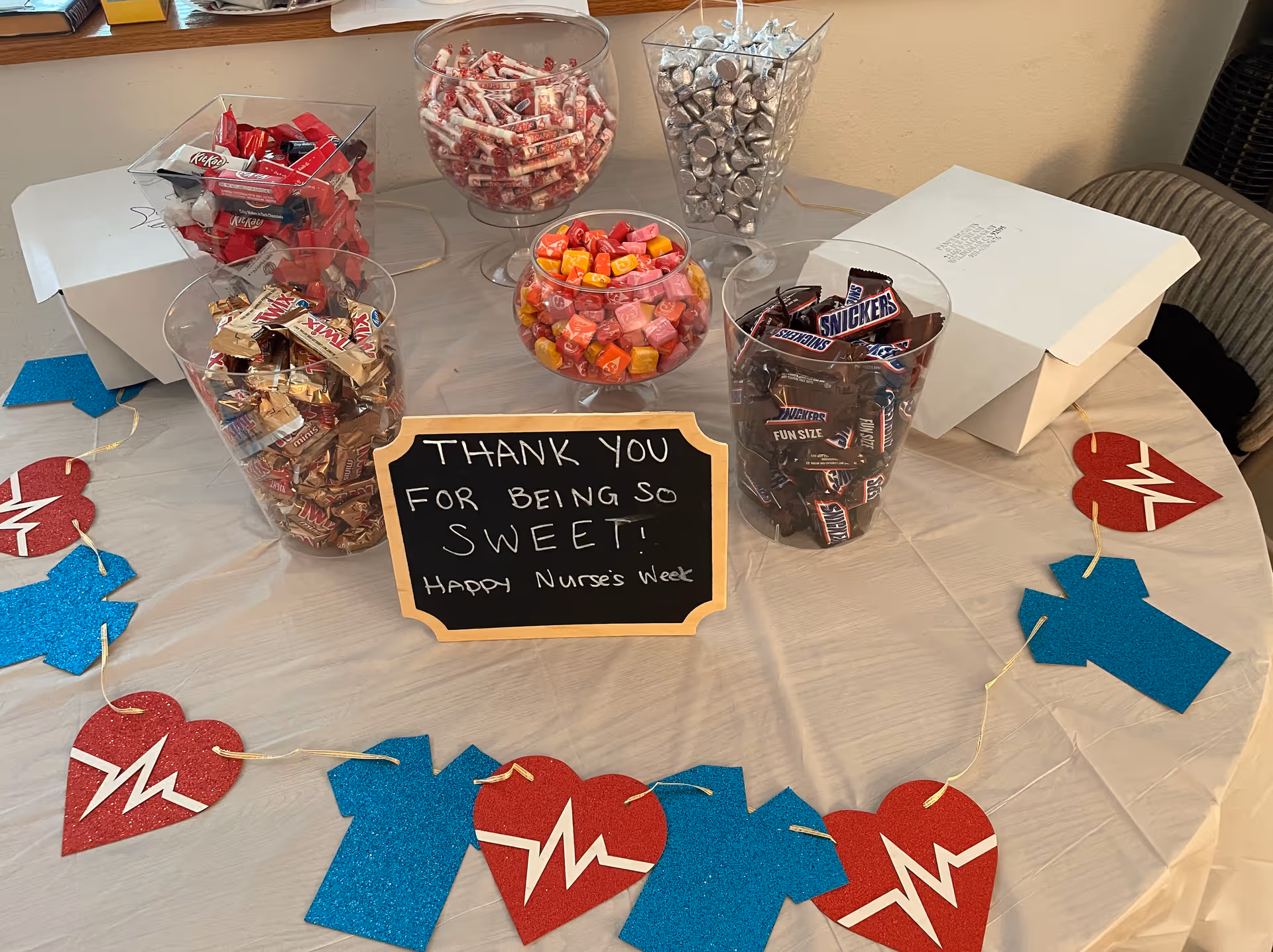 A table covered with a white tablecloth displaying several clear containers filled with various candies including Kit Kat, Smarties, Hershey's Kisses, Twix, and Snickers. In the center is a small chalkboard sign that reads 'Thank you for being so sweet! Happy Nurse's Week'. The table is decorated with a garland of red hearts with white heartbeat lines and blue nurse scrubs cutouts.