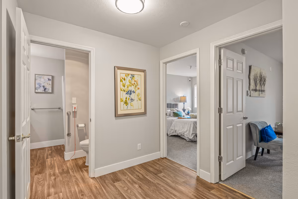 Interior hallway with wood flooring leading to three rooms: a bathroom with a toilet and grab bar, a bedroom with a bed and nightstand lamp, and a sitting area with a gray chair and blue pillow. A framed floral artwork hangs on the hallway wall.