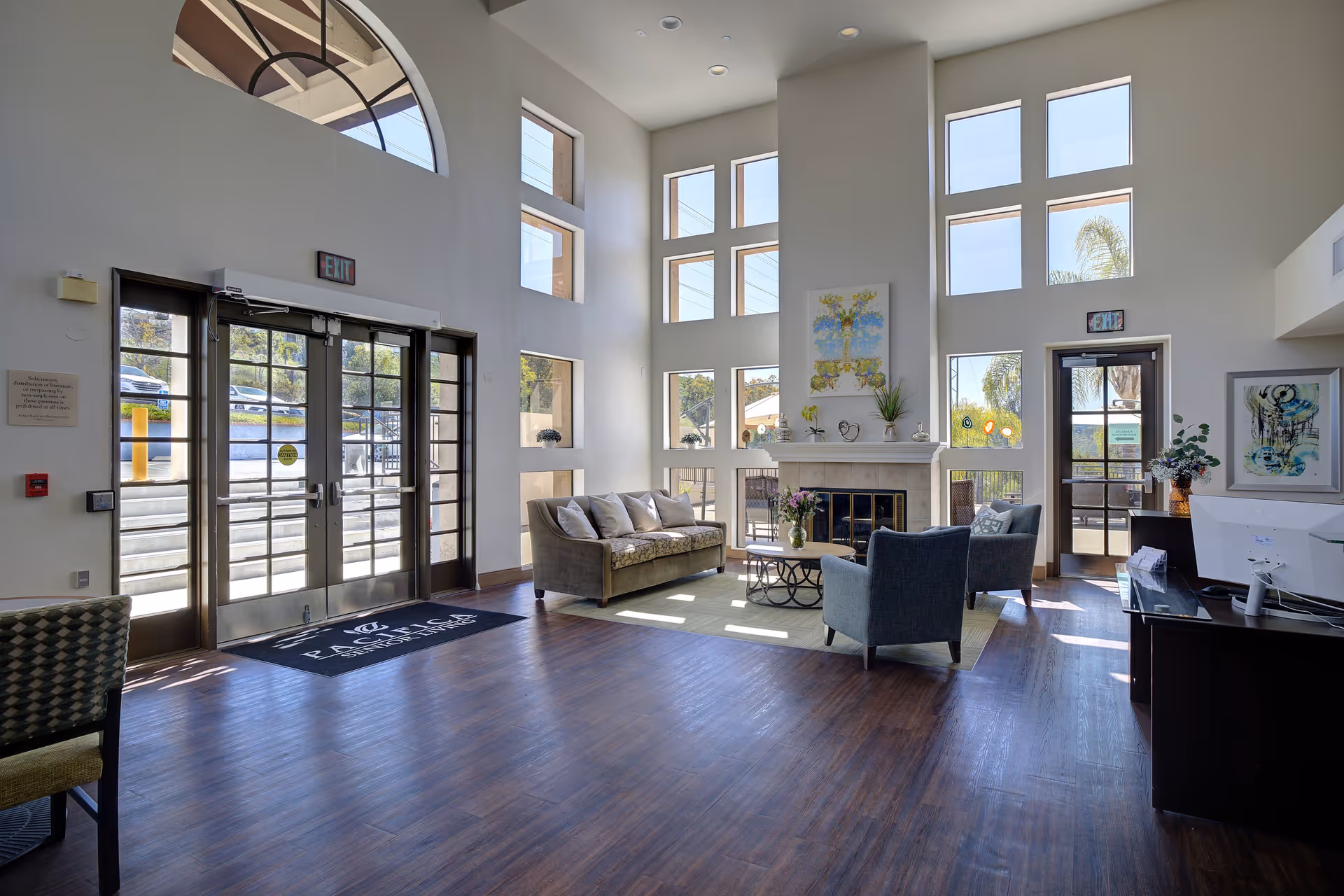 Bright and spacious senior living facility lobby with large windows letting in natural light, a seating area with a sofa and two armchairs around a coffee table, a fireplace with artwork above it, and double glass doors leading outside.