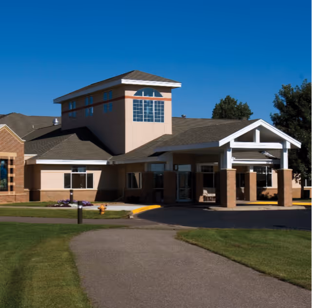 Front exterior of a senior living facility with a covered entrance, tower window feature, and surrounding lawn under a clear blue sky.