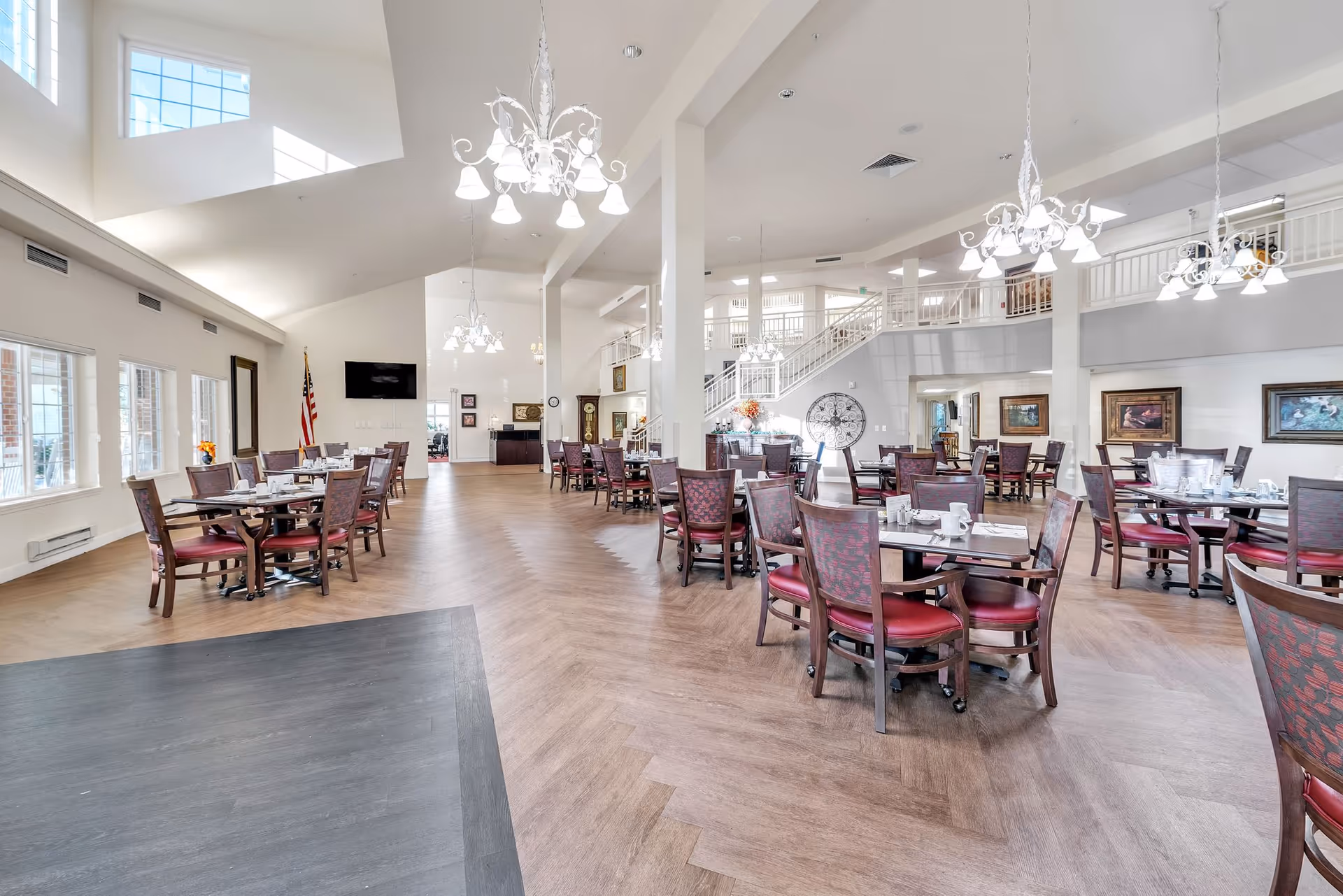 Spacious dining room with multiple round tables and chairs arranged neatly. The room features high ceilings with large windows allowing natural light to fill the space. Elegant chandeliers hang from the ceiling, and there are paintings on the walls. A staircase and an American flag are visible in the background.