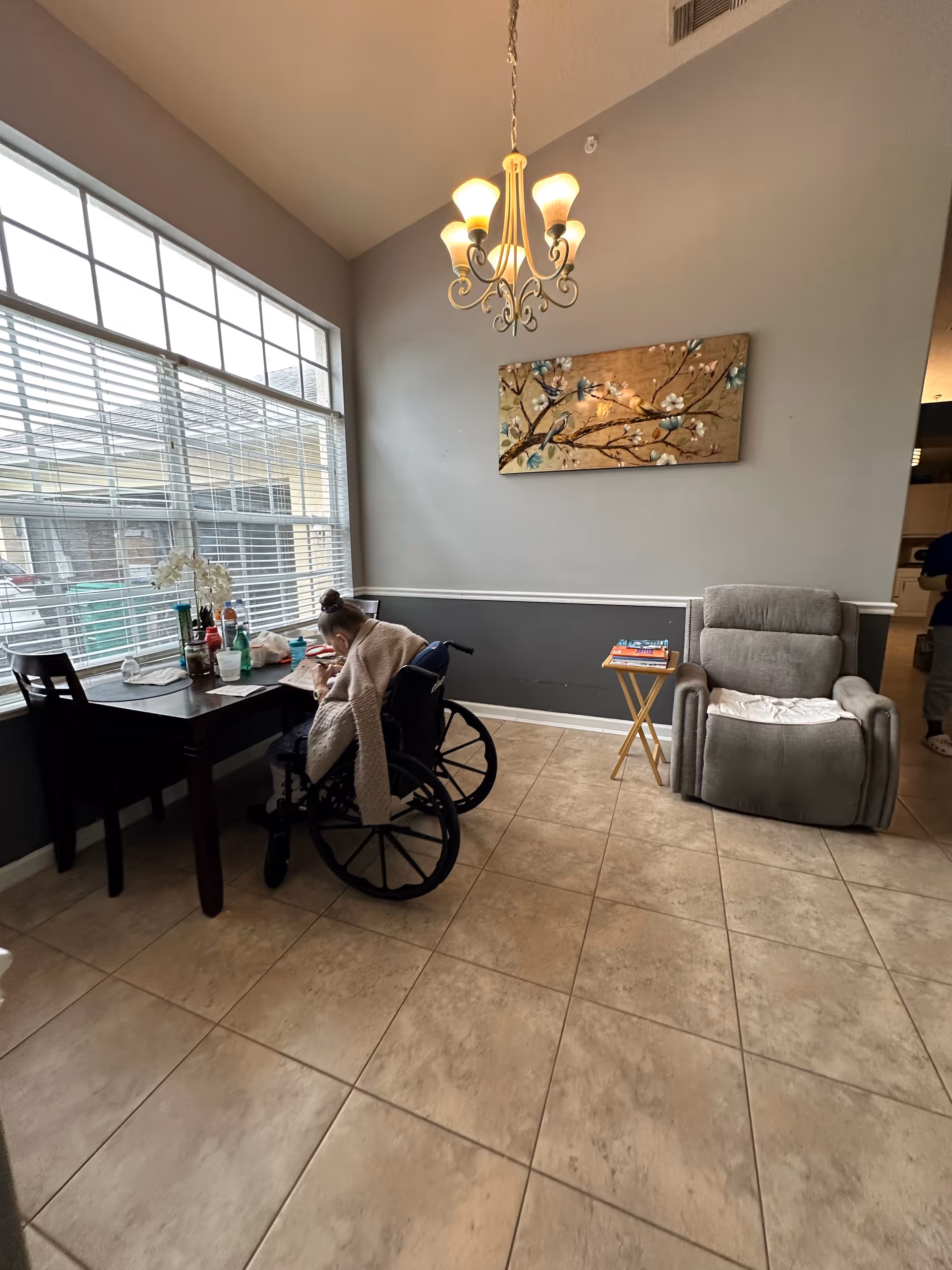 A person in a wheelchair sitting at a table near a large window with blinds, writing or drawing. The room has beige tiled floors, gray walls with white trim, a chandelier hanging from the ceiling, a floral painting on the wall, a gray recliner chair, and a small side table with books.
