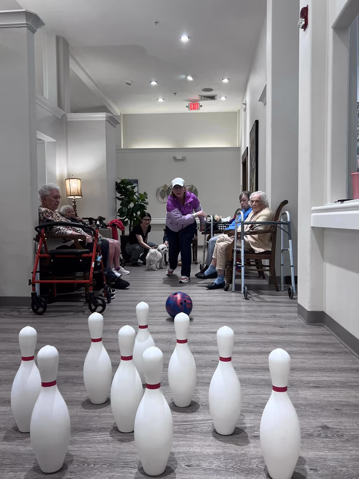 A group of elderly people and a caregiver are engaged in an indoor bowling game in a hallway. The elderly participants are seated or using walkers, watching as the caregiver rolls a bowling ball towards plastic pins set up on the floor. The setting appears to be a well-lit, spacious area with light-colored walls and wood flooring.