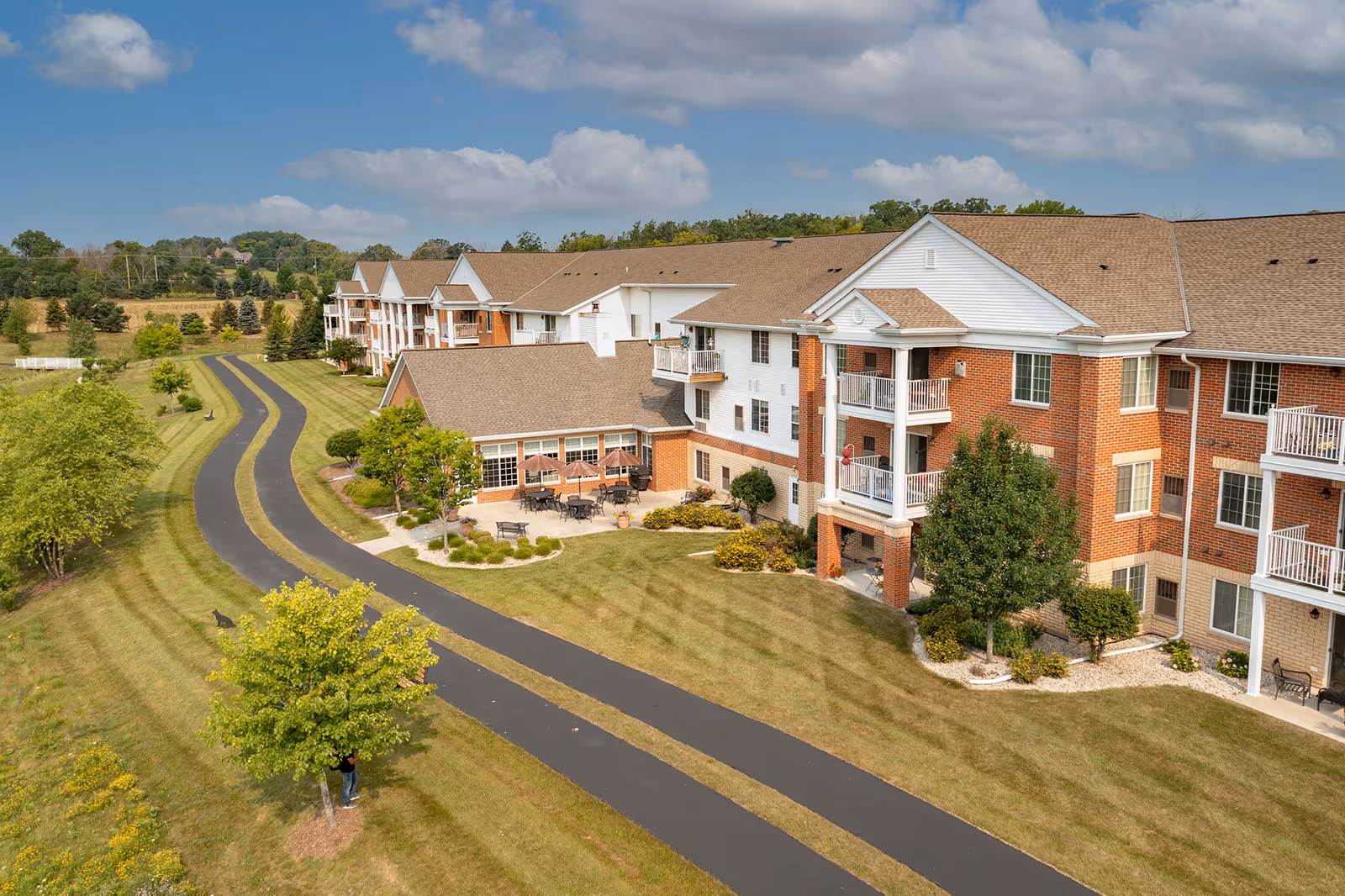 Exterior view of a multi-story senior living apartment building with brick and white siding. The building has balconies and a patio area with tables and umbrellas. A paved pathway winds through a well-maintained grassy area with trees and shrubs under a partly cloudy sky.