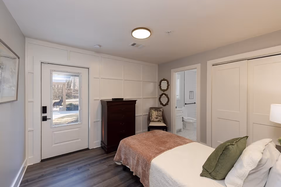 A cozy bedroom in a senior living facility featuring a bed with white and green pillows and a brown blanket. There is a dark wooden chest of drawers against a white paneled wall, a chair with a decorative cushion, and two small mirrors hanging above it. A door with a window leads outside, and another door opens to a bathroom with a visible toilet and grab bars. The room has wood flooring and neutral-colored walls.