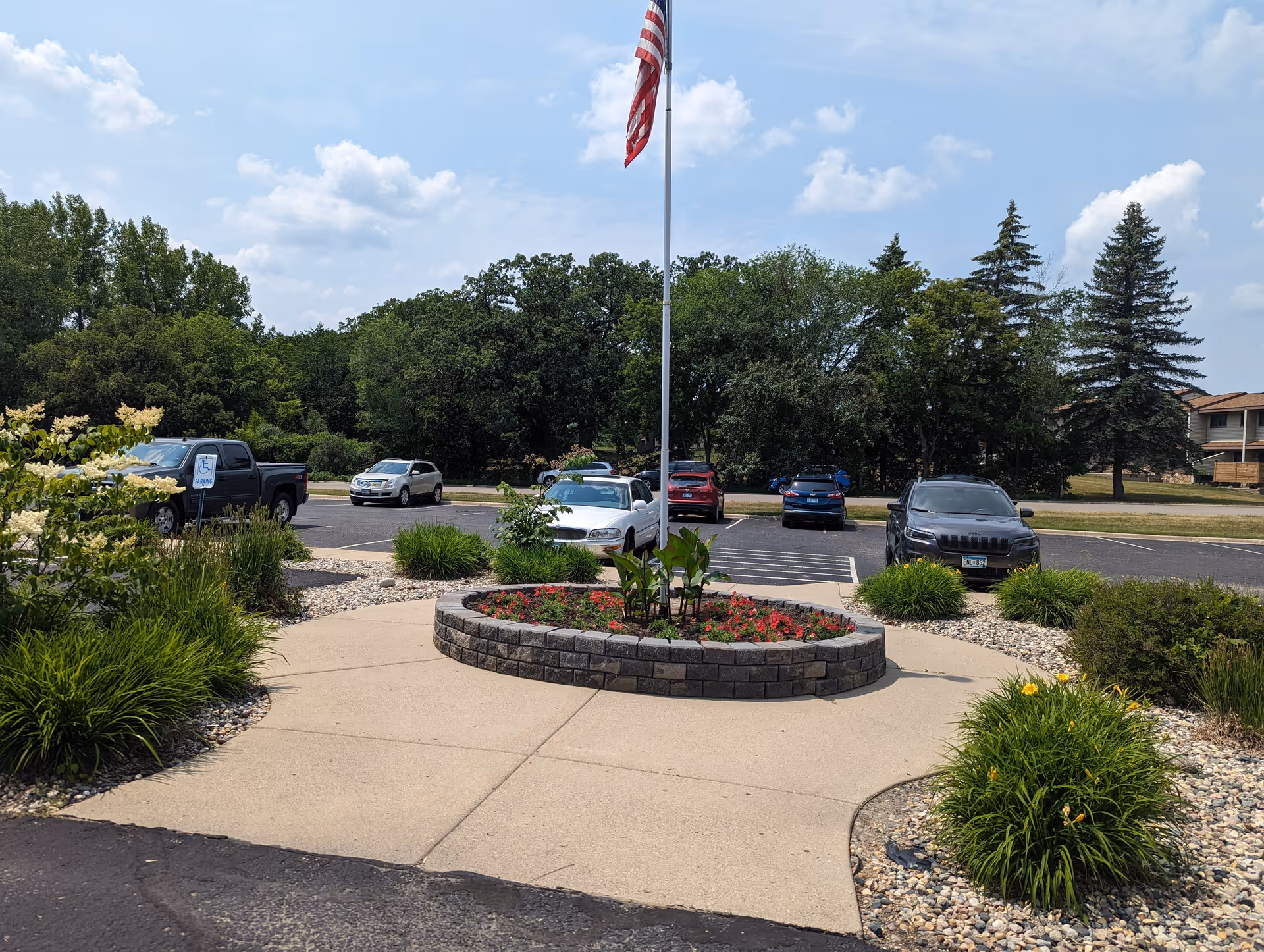 Landscaped front entrance with a circular raised flower bed, an American flag on a pole, and a parking lot with several cars.