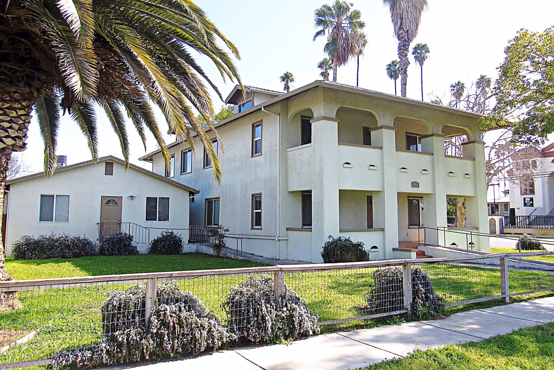 Exterior view of a two-story beige building with an arched balcony and a smaller adjacent building, surrounded by a green lawn, palm trees, and a wire fence along the sidewalk.