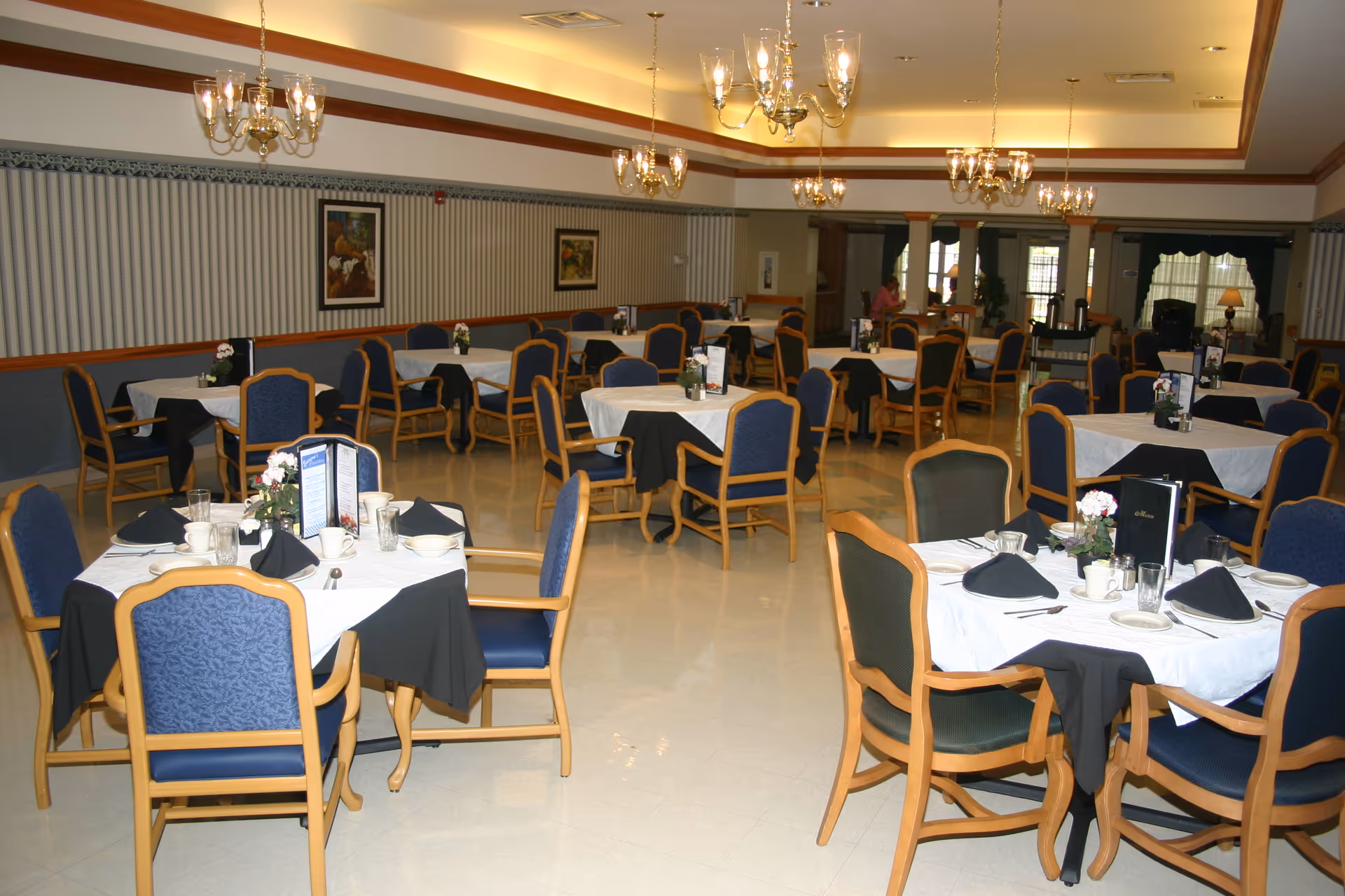 A spacious dining room with multiple round tables covered with white tablecloths and black napkins. Each table is set with plates, cups, glasses, silverware, and small flower arrangements. The room has blue cushioned chairs with wooden frames, chandeliers hanging from the ceiling, and framed artwork on the walls.