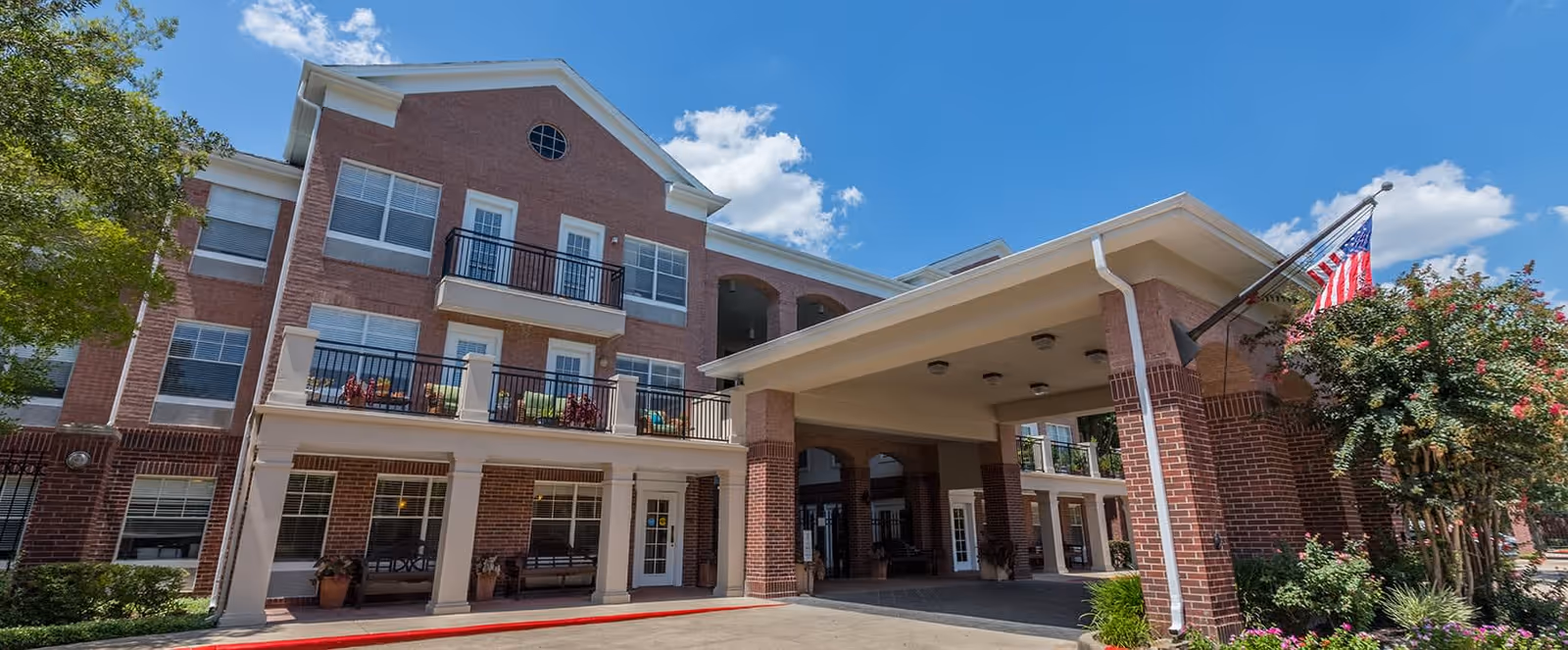 Exterior view of a multi-story brick building with balconies and large windows under a blue sky with some clouds. The building has a covered entrance with columns and an American flag mounted on the right side near some flowering bushes.