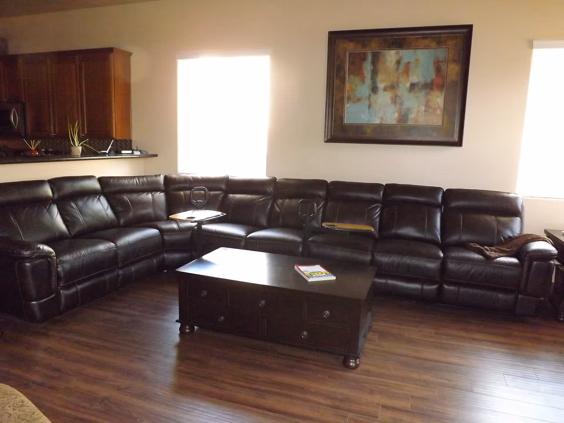 A living room with a large dark brown leather sectional sofa arranged in an L-shape. There is a dark wooden coffee table in front of the sofa with a book on it. The room has wooden flooring and two windows with blinds partially drawn. A colorful abstract painting is hung on the wall above the sofa. In the background, part of a kitchen with wooden cabinets and a countertop is visible.