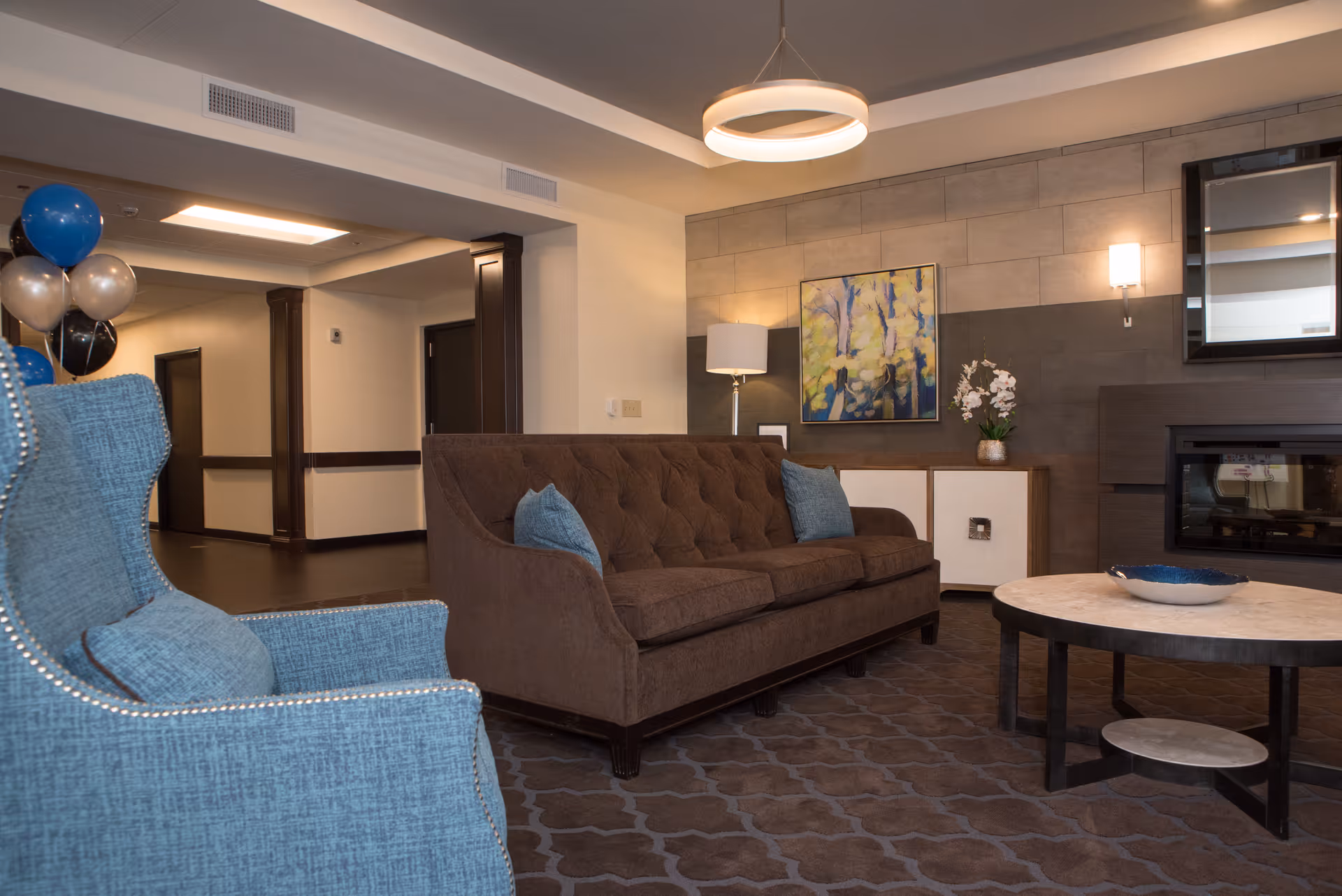A modern lounge area with a brown tufted sofa, blue armchairs, coffee table and wall art in a senior living facility.