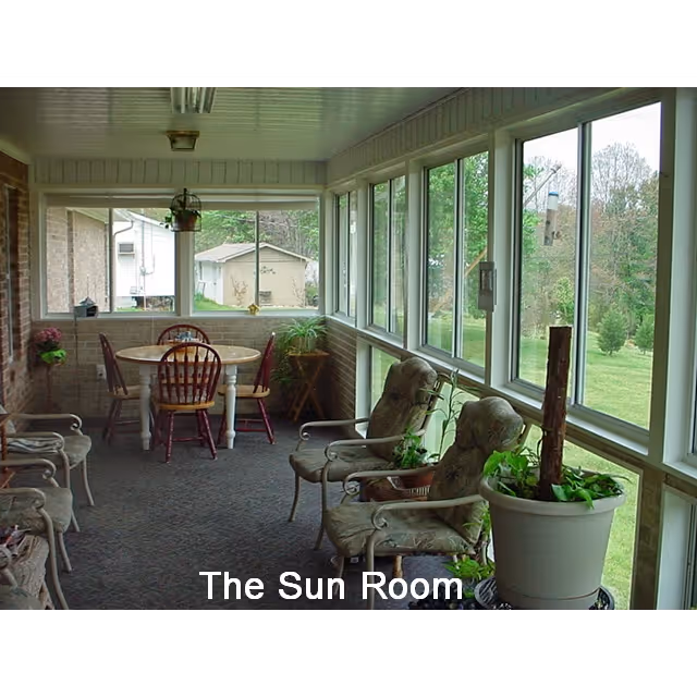 A sunroom with large windows on two sides letting in natural light, featuring a round wooden table with four chairs, several cushioned armchairs, and various potted plants. Outside, a green lawn and trees are visible.