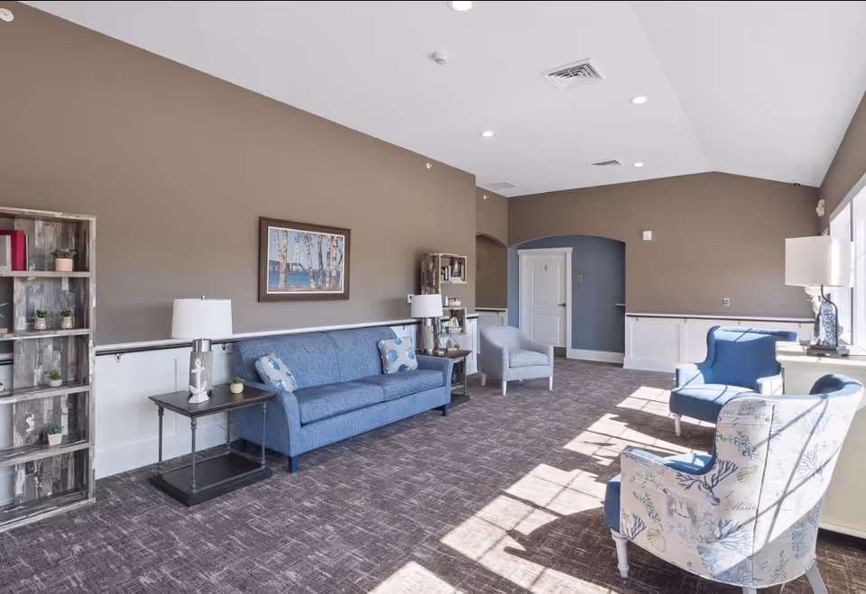 A bright and spacious living room area with a blue sofa, two patterned armchairs, a gray armchair, two side tables with lamps, a wooden bookshelf with small plants and decor, and a framed picture on the wall. The room has beige walls, carpeted floor, and large windows letting in natural light.