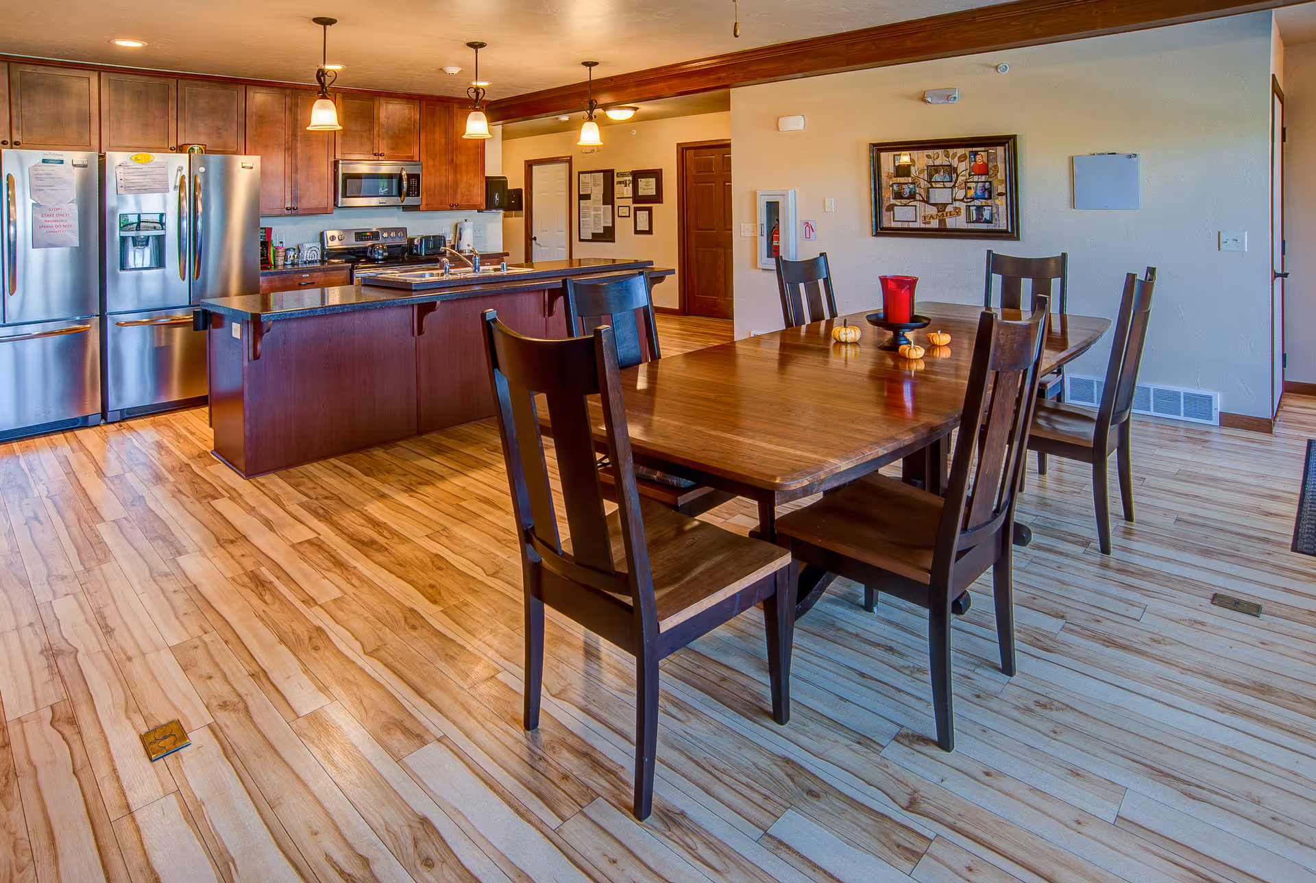 A spacious kitchen and dining area with wooden flooring and cabinetry. The kitchen features stainless steel appliances including two refrigerators, a microwave, and an oven. A large island with a dark countertop and a sink is in the center. The dining area has a wooden table with six chairs, decorated with small pumpkins and a red candle holder. Warm lighting fixtures hang from the ceiling, and framed pictures and notices are visible on the walls.