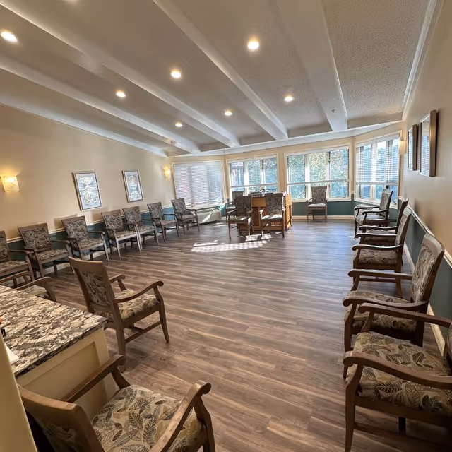 A spacious, well-lit room with wooden flooring and a high ceiling featuring exposed beams. The room is furnished with multiple cushioned chairs arranged along the walls and around a small table near large windows that let in natural light. The walls are decorated with framed artwork and wall-mounted lights.