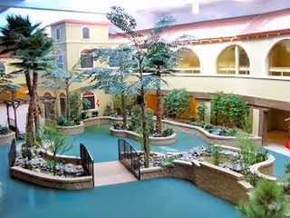 Indoor courtyard area with a variety of plants and trees, surrounded by a two-story building with arched windows and a red roof. The courtyard features raised garden beds with greenery and a small bridge over a pathway.