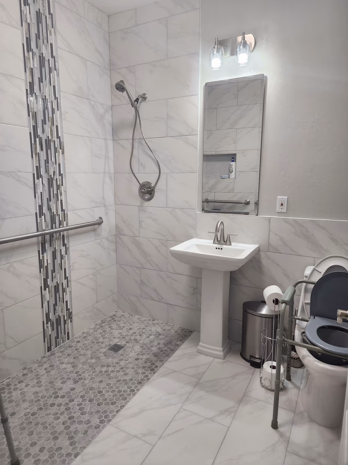 A modern bathroom with white and gray marble tiles. The walk-in shower has a handheld showerhead and a vertical strip of decorative tiles. There is a white pedestal sink with a mirror and light fixture above it. A toilet with a raised seat and safety rails is visible, along with a trash can and toilet paper holder.
