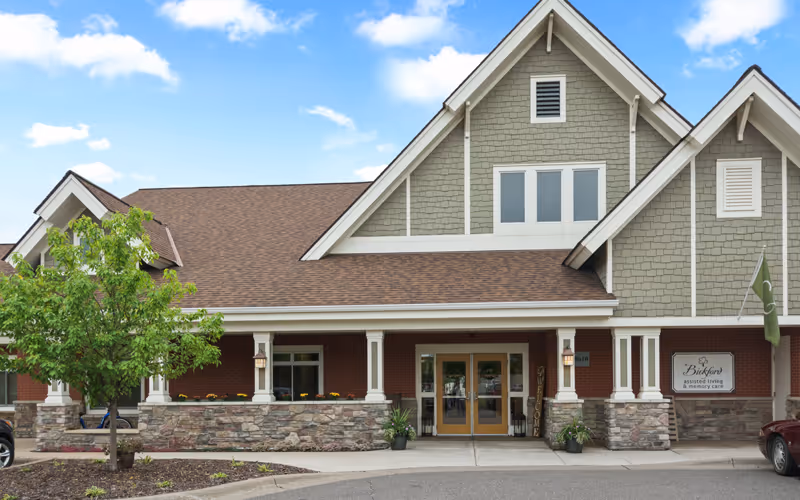 Front entrance of a two-story assisted living facility with a covered porch, double glass doors, peaked roof, and landscaping.