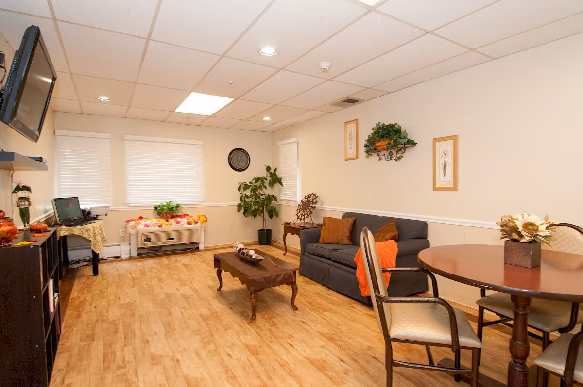 A cozy living room with a wooden floor, a gray sofa with orange and brown pillows, a wooden coffee table, a round dining table with chairs, a wall-mounted TV, a computer desk, and several plants and framed pictures on the walls.