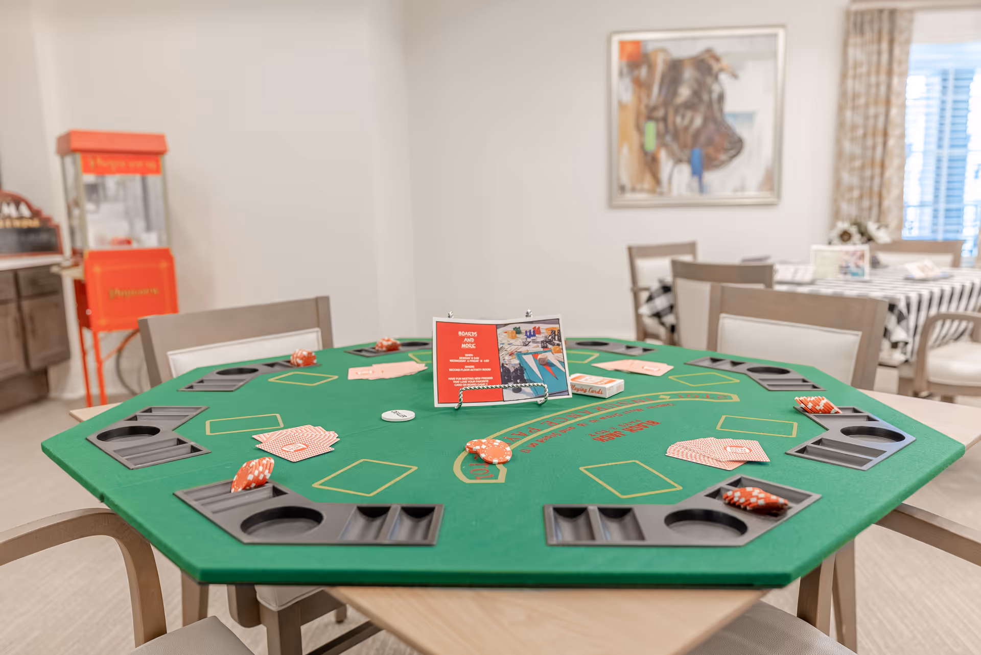 A green octagonal poker table set up with playing cards and poker chips in a well-lit room. The table has built-in cup holders and card slots. In the background, there is a red popcorn machine, a painting of a dog on the wall, and another table with a black and white checkered tablecloth.