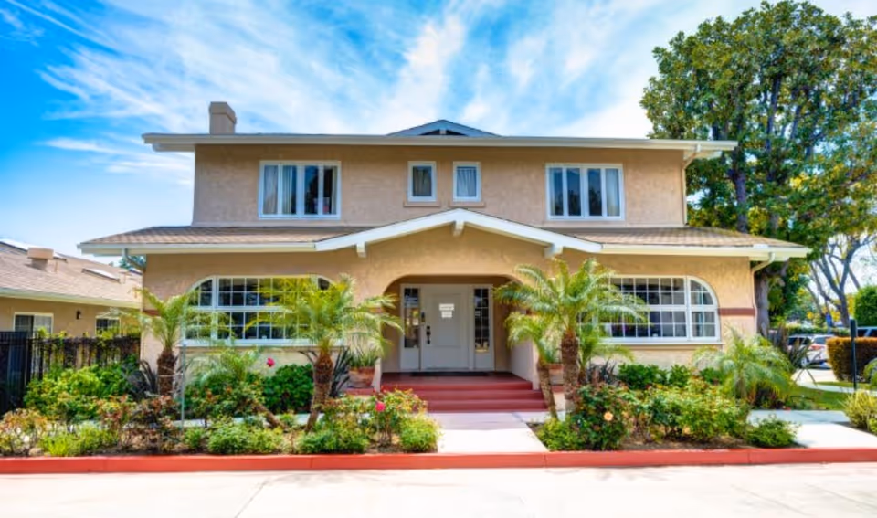 Two-story beige memory care facility entrance with red steps and lush tropical landscaping.
