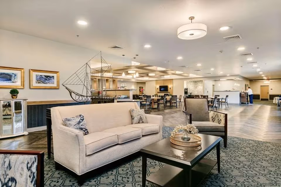 A spacious and well-lit senior living common area with a beige sofa and an armchair around a dark coffee table on a patterned rug. In the background, there are multiple dining tables and chairs, a decorative metal sailboat sculpture, framed artwork on the wall, and a person standing near a counter.