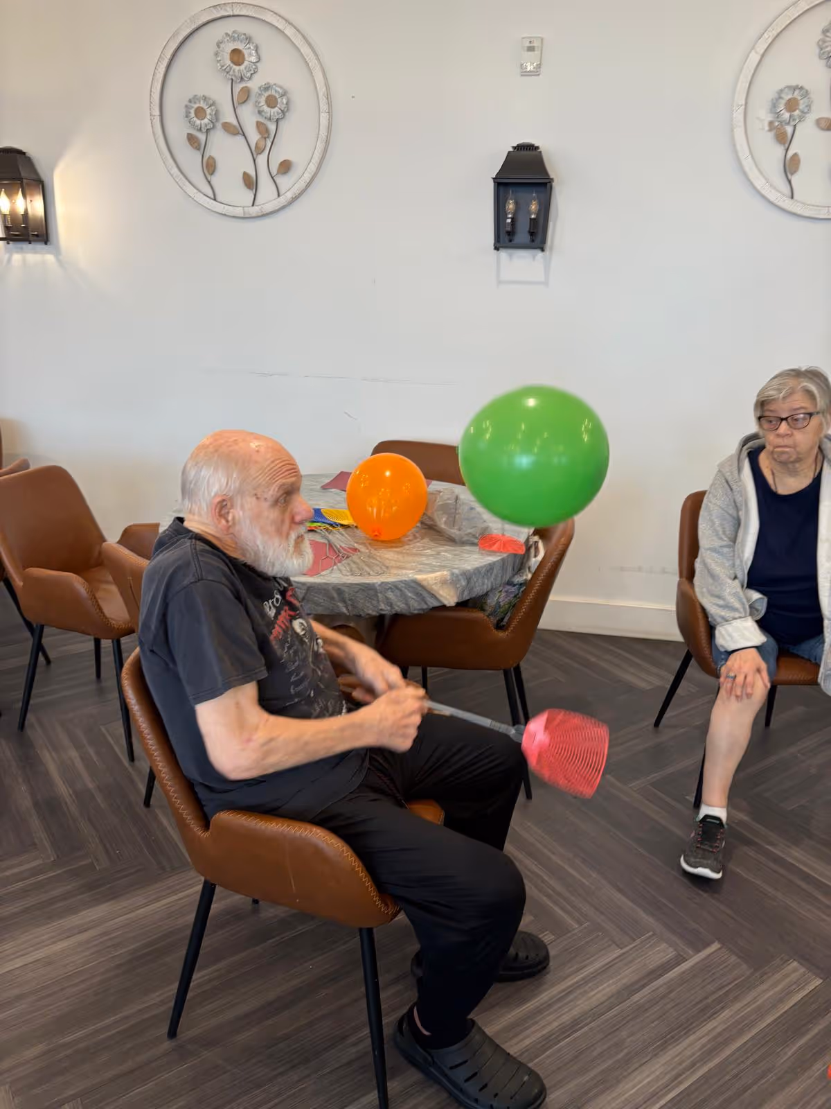 An elderly man and woman seated in a common room with brown chairs and a table covered with a plastic tablecloth. The man is playing with a green balloon using a pink fly swatter, while the woman watches. The room has light-colored walls decorated with round floral wall art and black wall sconces.