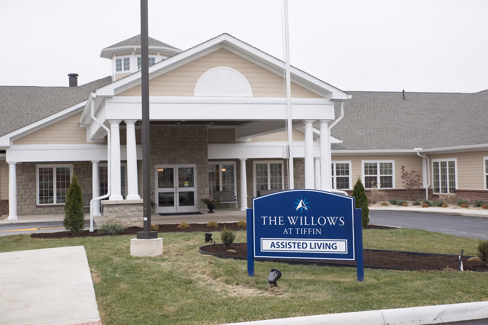 Exterior front view of The Willows at Tiffin assisted living facility showing the entrance with white columns, beige siding, stone accents, and a blue sign with the facility name and 'Assisted Living' on the lawn.