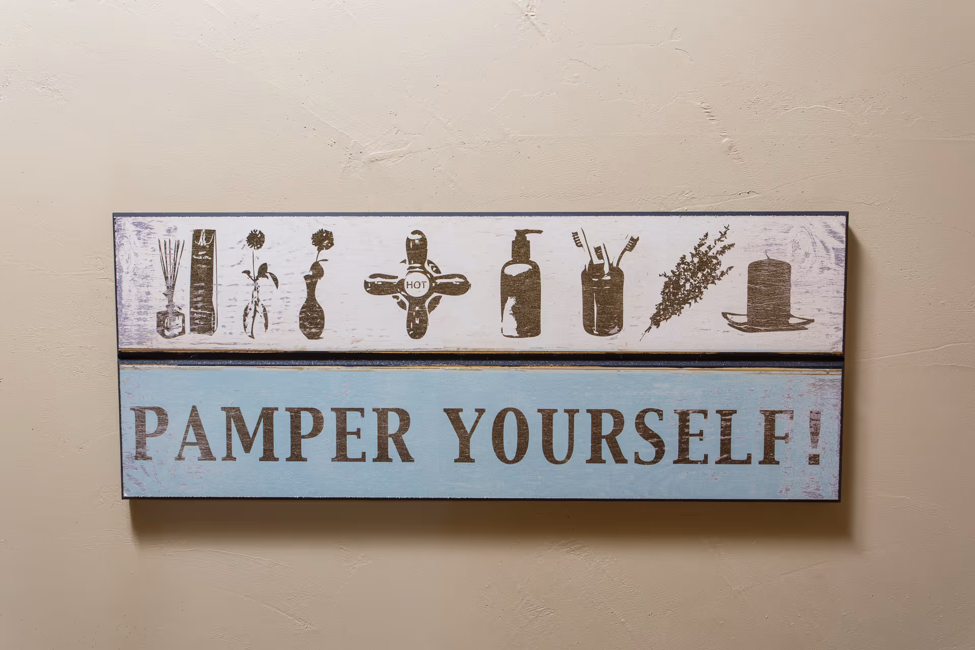A decorative wall sign reading 'PAMPER YOURSELF!' with spa-related icons mounted on a beige wall.