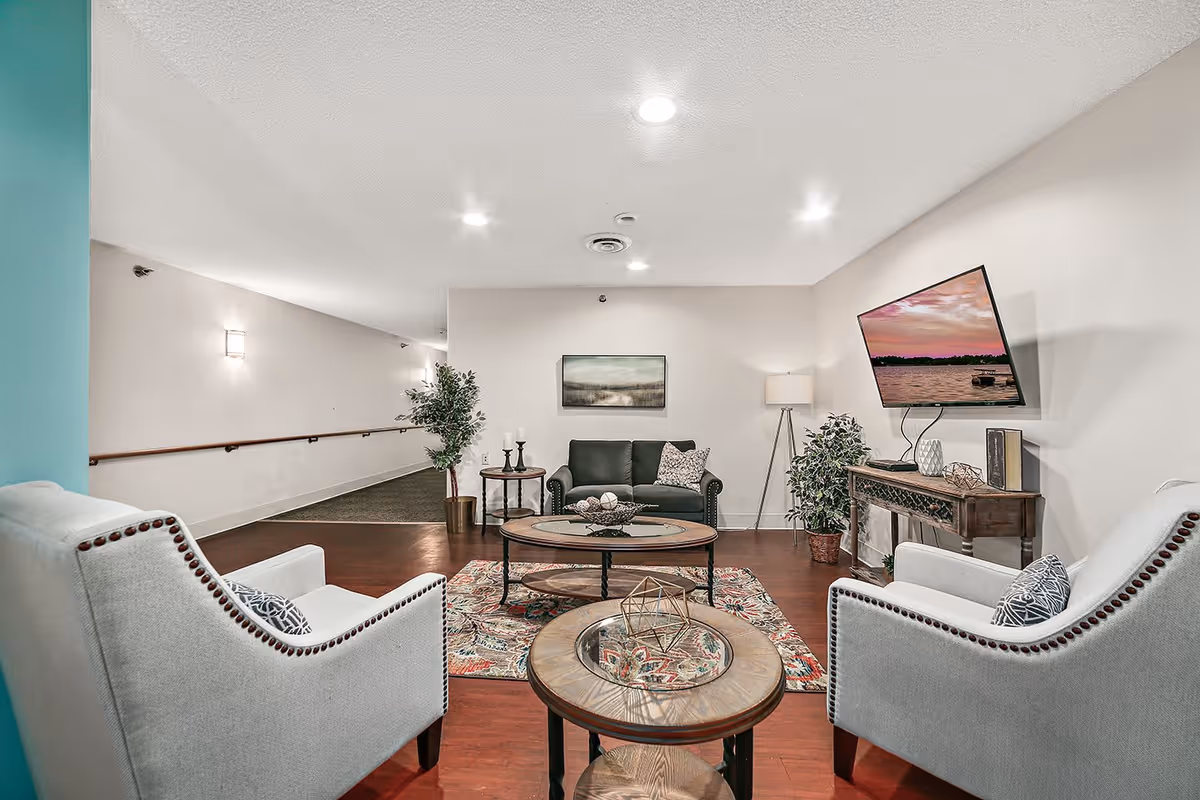 A cozy living room area with two light gray armchairs facing a dark green loveseat. There are two round wooden coffee tables with decorative items on them, a colorful patterned rug, a wooden console table with decor, a floor lamp, and a wall-mounted TV displaying a sunset over water. The walls are light-colored, and there is a hallway with handrails visible in the background.