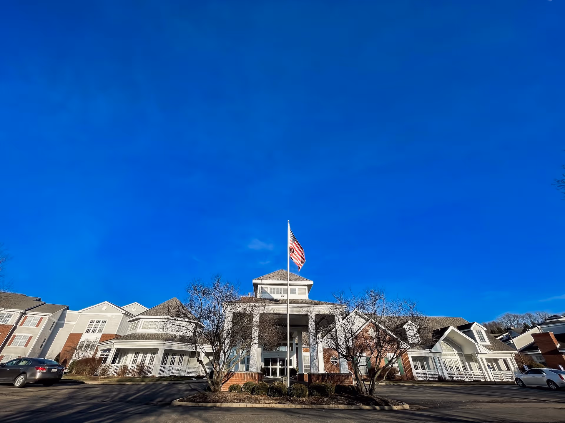 Front exterior of a senior living community building with an American flag on a flagpole under a clear blue sky.