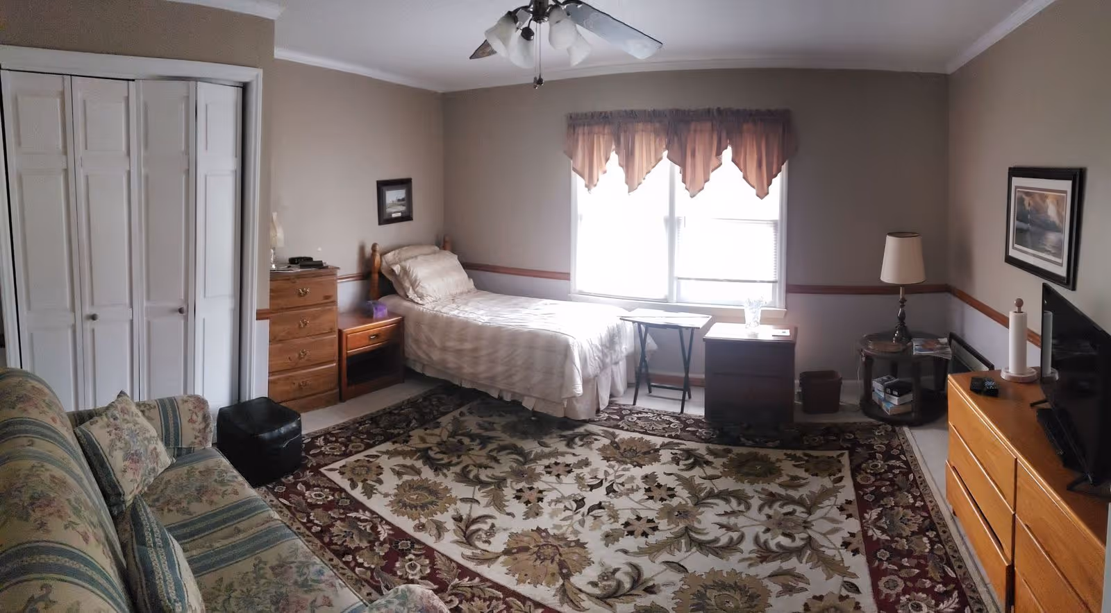 A cozy bedroom in an assisted living community featuring a single bed with beige bedding, a floral patterned sofa, a large decorative rug with floral designs, a wooden dresser with a TV on top, a small wooden chest of drawers, a nightstand, a side table with a lamp, and a window with sheer curtains letting in natural light.