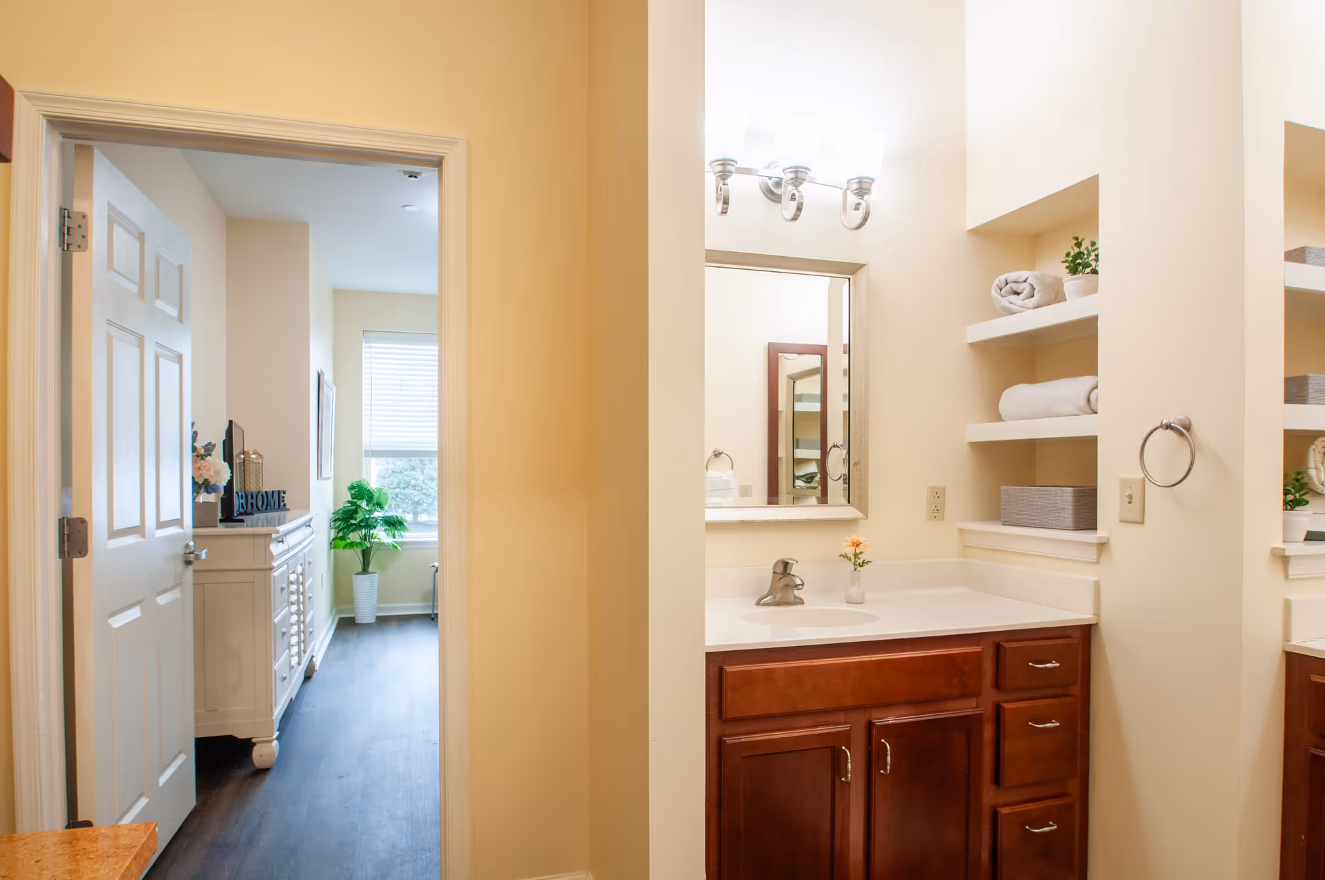 View of a bathroom vanity with a sink, mirror, and shelves holding towels and a small plant. To the left, an open door leads to a bright room with a window, a white dresser, and a potted plant.