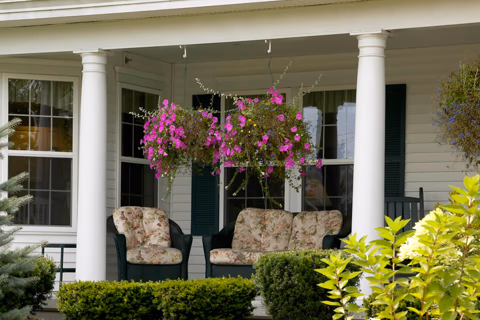 A porch area with white columns, floral cushioned wicker chairs, hanging baskets of vibrant pink flowers, and green shrubs in the foreground.