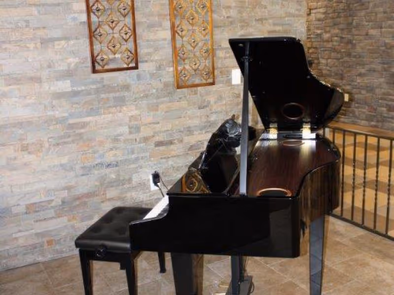 A black grand piano with its lid open is placed in a room with stone tile walls and floor. There is a black cushioned piano bench in front of the piano. Two decorative wooden wall hangings are mounted on the wall behind the piano.