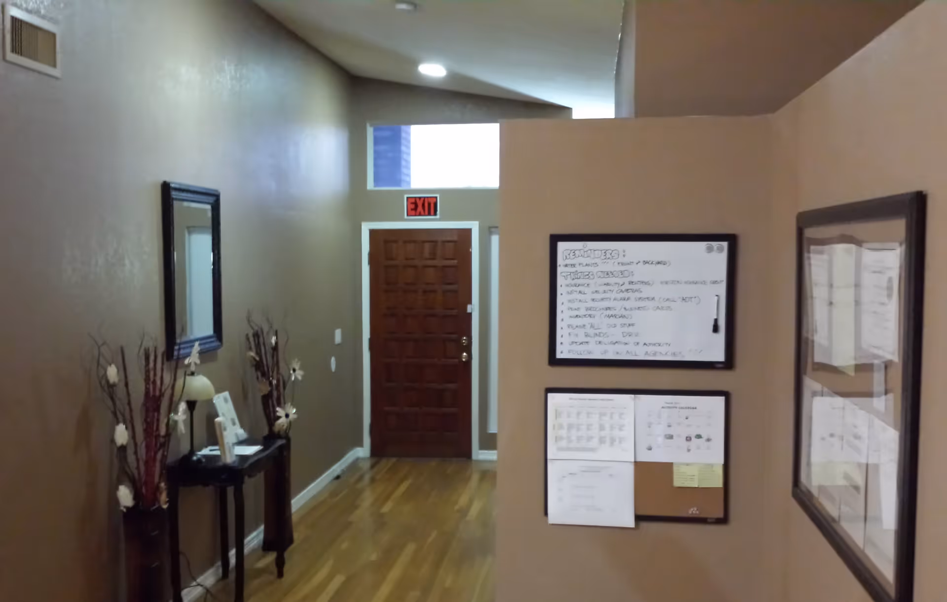 Interior hallway of a senior living facility with a wooden front door at the end, an exit sign above the door, a small table with a lamp and decorative branches on the left, and two bulletin boards with notices on the right wall.