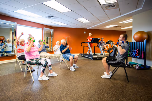 A group of elderly people seated on chairs in a fitness room participating in a guided exercise session using small dumbbells. A fitness instructor is leading the session, also seated and holding dumbbells. The room has mirrors on one wall, exercise equipment including treadmills and stationary bikes, and a rack with various weights and exercise balls.