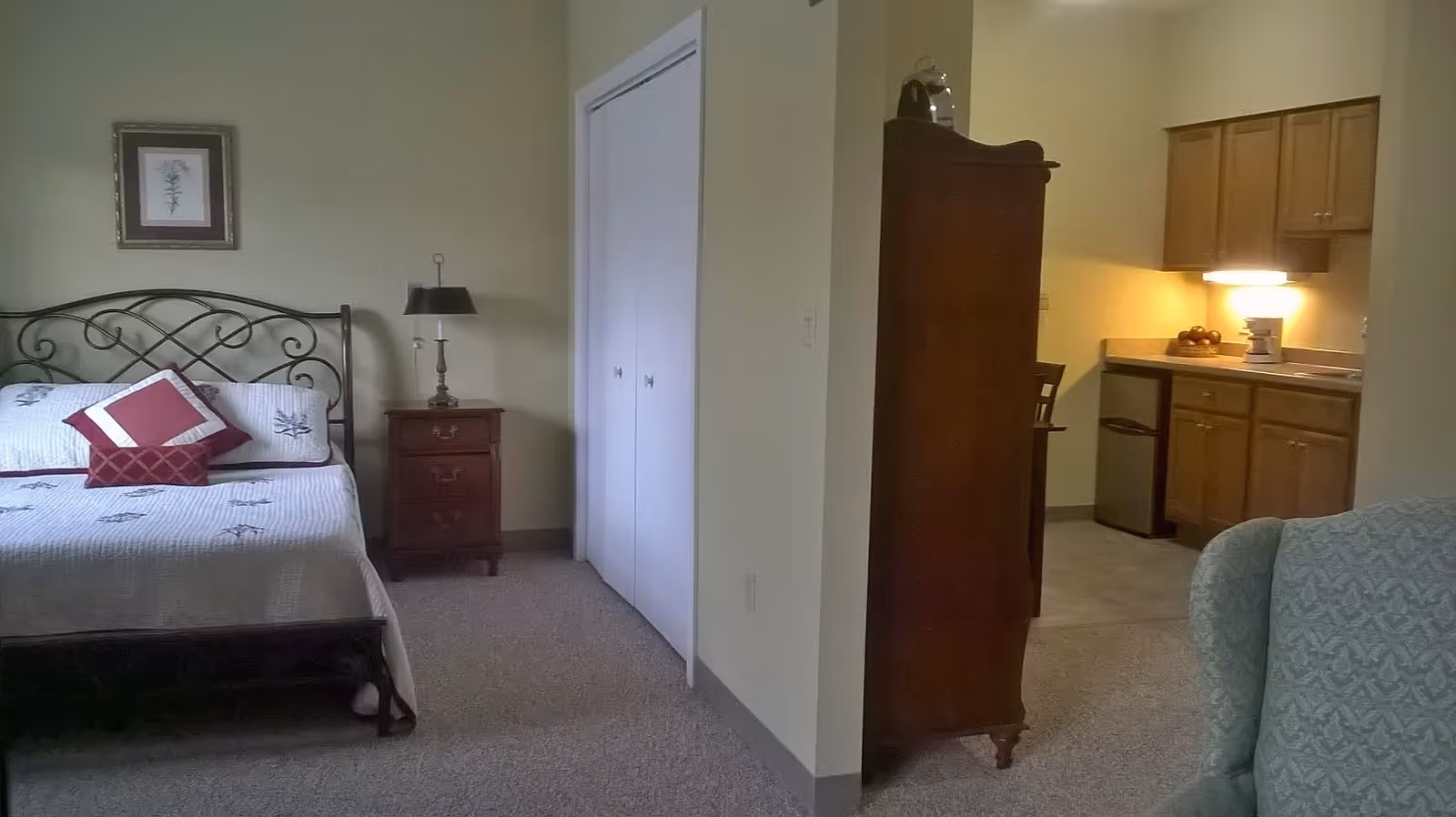 Interior view of a senior living facility room at Heritage Woods showing a bed with decorative pillows, a nightstand with a lamp, a framed picture on the wall, a closet with white double doors, a wooden cabinet, a small kitchen area with wooden cabinets, a mini fridge, and a countertop with a lamp and some items, and a patterned armchair partially visible in the foreground.