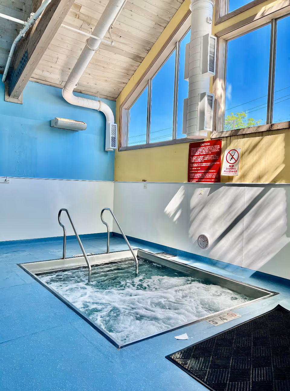 Indoor hot tub with bubbling water, metal handrails, blue flooring, and large windows letting in natural light. The walls are painted blue and yellow, with ventilation ducts visible near the ceiling. Safety signs are posted on the wall.