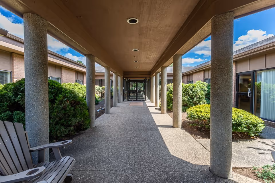 Covered walkway with concrete pillars leading to glass double doors of a building, surrounded by green bushes and outdoor chairs on the left side under a partly cloudy blue sky.