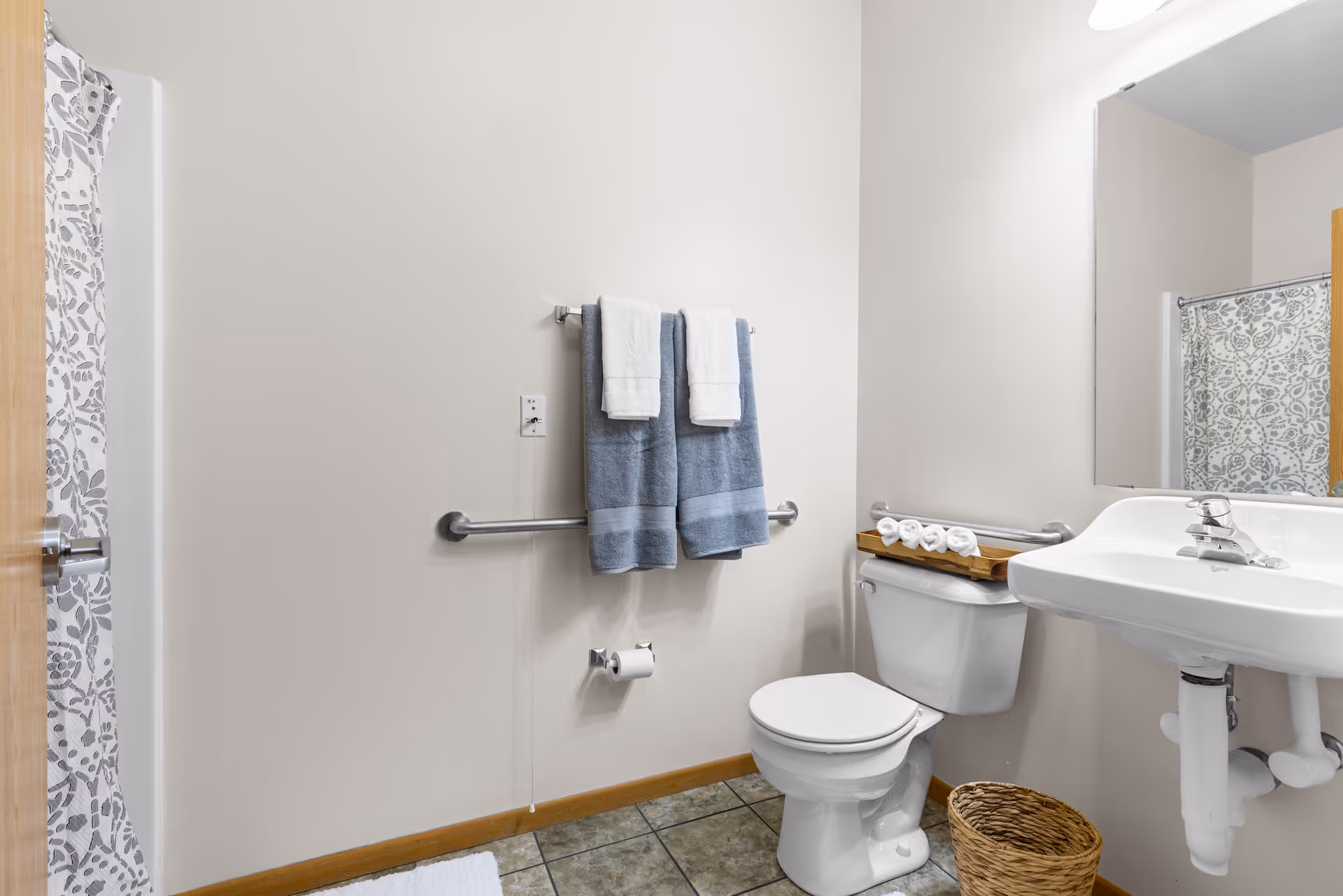 Bright accessible bathroom featuring a toilet with grab bars, a pedestal sink, towel rack, and a shower with patterned curtain.
