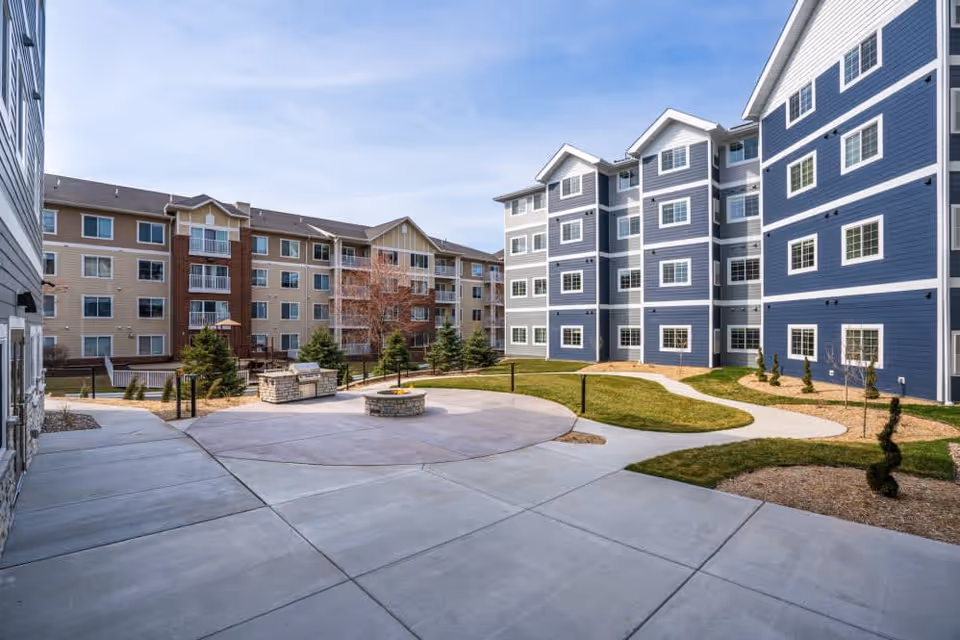 Outdoor courtyard area of Hayden Grove Bloomington featuring a circular concrete patio with a fire pit in the center, surrounded by pathways, grass, small trees, and multi-story residential buildings in the background under a clear sky.