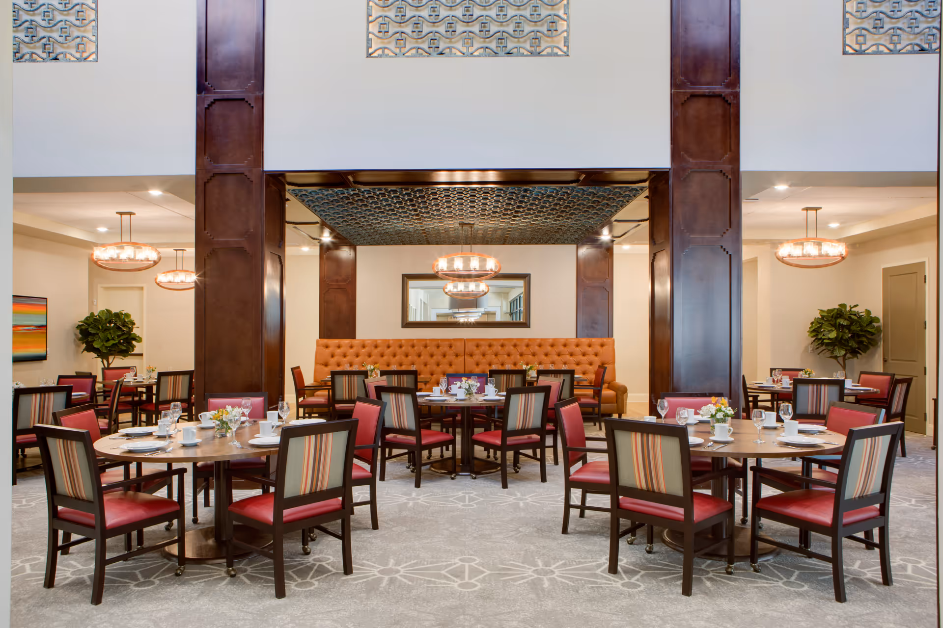 A spacious dining room with multiple round wooden tables set with white plates, cups, and glassware. Each table is surrounded by chairs with red seats and striped backs. The room features tall dark wooden pillars, a patterned ceiling section above the central area, and modern chandeliers. There are plants and colorful artwork on the walls, and a long tufted orange bench along the back wall.