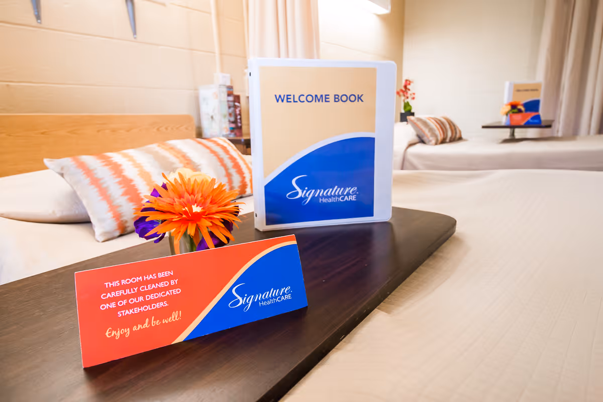 Two beds in a clean, well-lit room with patterned pillows and a dark wooden tray on one bed holding a small vase with orange and purple flowers, a welcome book, and a card indicating the room has been carefully cleaned by dedicated stakeholders.