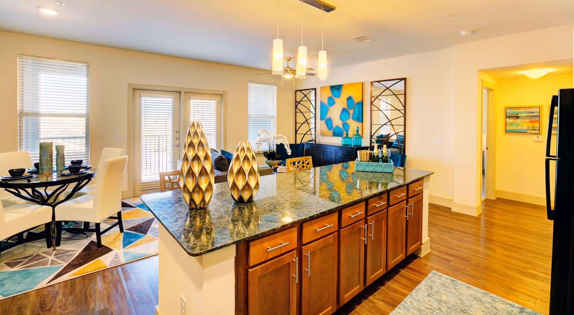 Bright and modern open-concept kitchen and dining area with a large granite island countertop featuring decorative vases. The dining area has a round table with white chairs on a colorful geometric rug. In the background, there is a living space with a blue and yellow abstract painting and large windows with blinds letting in natural light.