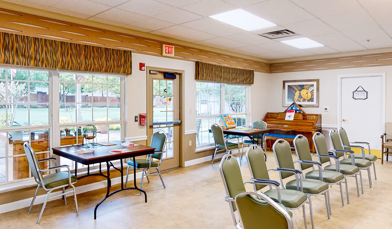 A bright room with large windows showing a green outdoor area. Inside, there are two tables with chairs around them, and a row of green cushioned chairs with metal armrests arranged in a line. A wooden piano with a folded American flag and a framed picture above it is against the wall. The room has beige walls, a light-colored floor, and a ceiling with recessed lighting. There is an exit door with a red exit sign above it and a white door with a welcome sign on the right side.