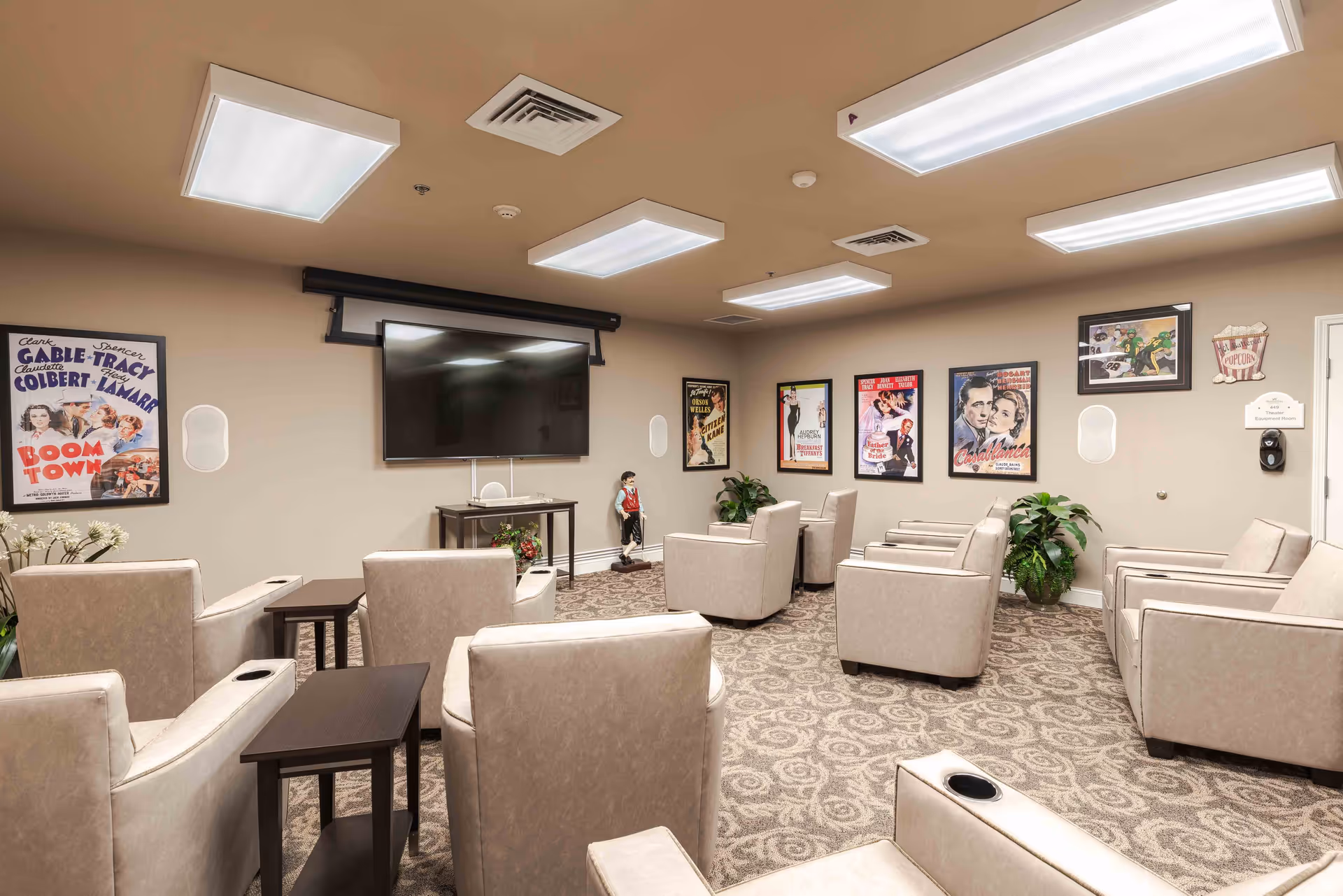A cozy theater room with beige armchairs arranged in rows facing a large flat-screen TV mounted on the wall. The walls are decorated with framed classic movie posters and there are potted plants in the corners. The room has patterned carpet flooring and bright ceiling lights.