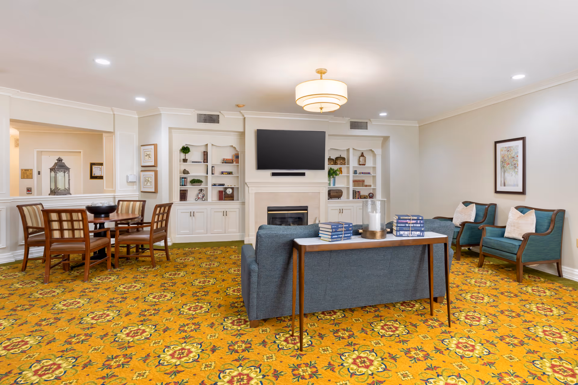 A cozy living room area with a patterned yellow carpet, a blue sofa with a wooden console table behind it holding stacks of books and a candle. Two blue armchairs with white pillows are positioned against the wall with a framed artwork above. A fireplace with built-in white shelves filled with books and decorative items is centered on the far wall, topped by a mounted flat-screen TV. A round wooden table with four chairs is visible to the left near an open doorway.