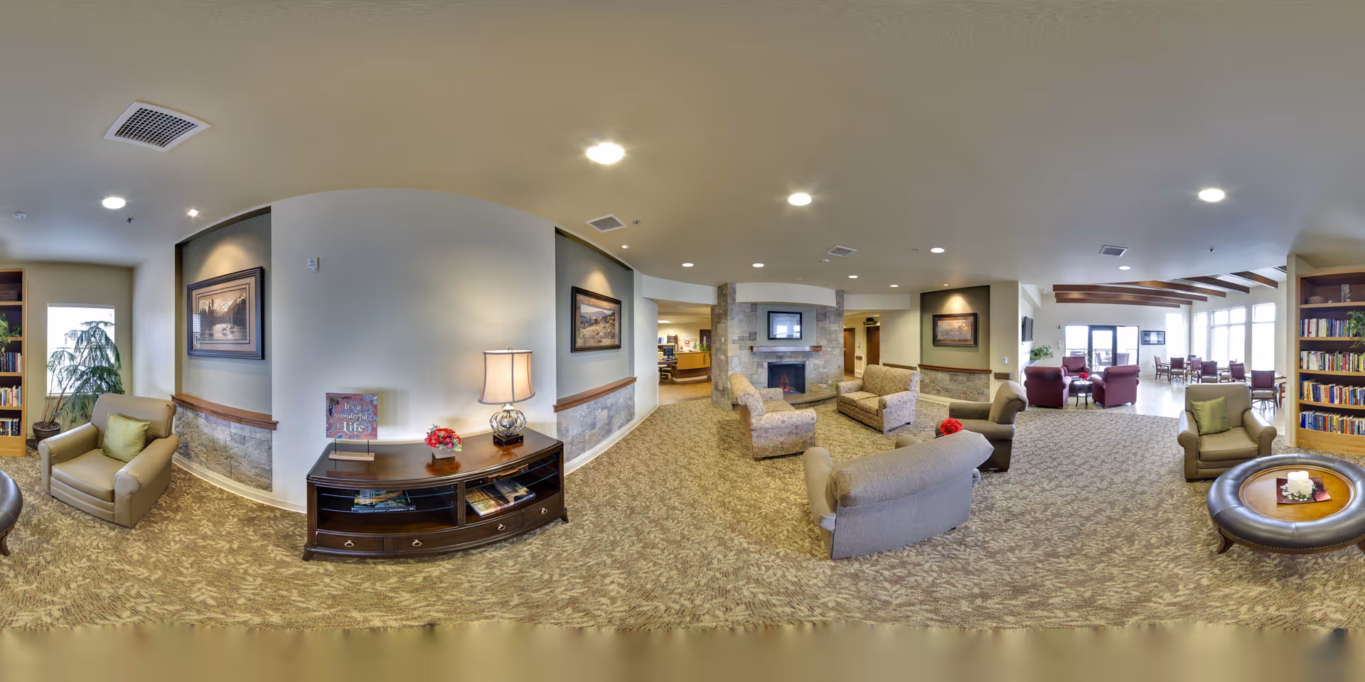 Spacious senior living lounge with sofas and armchairs arranged around a fireplace, bookshelves, and a dining area by large windows.